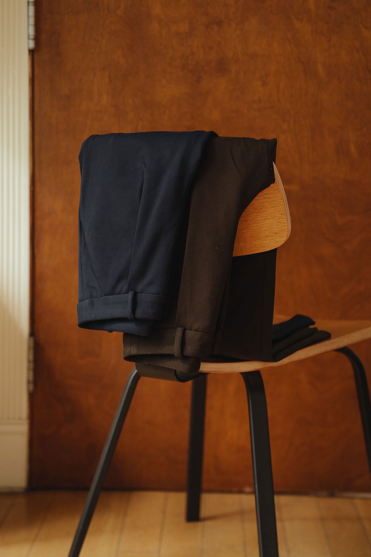 Petite dress pants in Espresso brown and Midnight navy blue, one pair of each colour draped over a chair, The Petite Standard